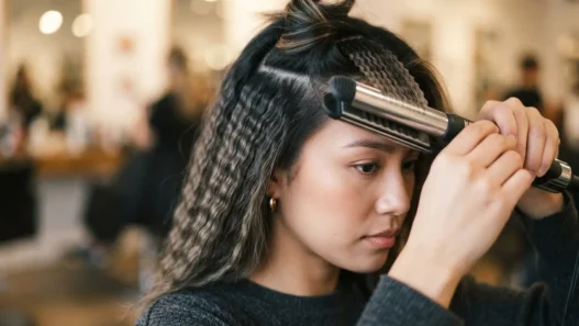 Woman closing a crimping iron on a lifted brunette section with already-crimped waves visible beside it in warm studio light