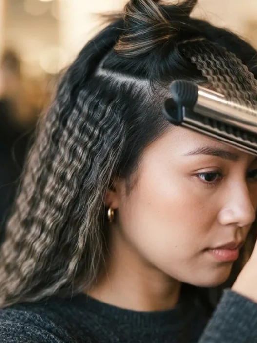 Woman closing a crimping iron on a lifted brunette section with already-crimped waves visible beside it in warm studio light