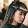 Woman closing a crimping iron on a lifted brunette section with already-crimped waves visible beside it in warm studio light