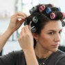 Woman placing magnetic rollers in the crown row of her dark hair, forward rolling direction visible, pinning as she works