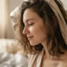Woman in satin sleep wrap half undone in soft morning window light, side profile from wrap to collarbone