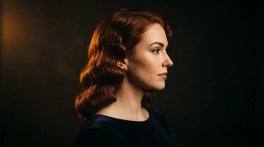 Woman with deep copper-auburn vintage red hair in set waves lit by warm amber gel light against black background